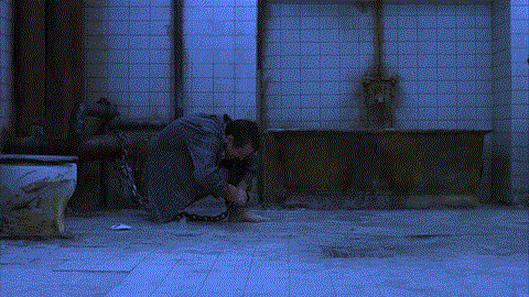 Adam tries to remove his shackles. He is bathed in fluorescent blue light, making the white tiles and himself take on a bluish tone.